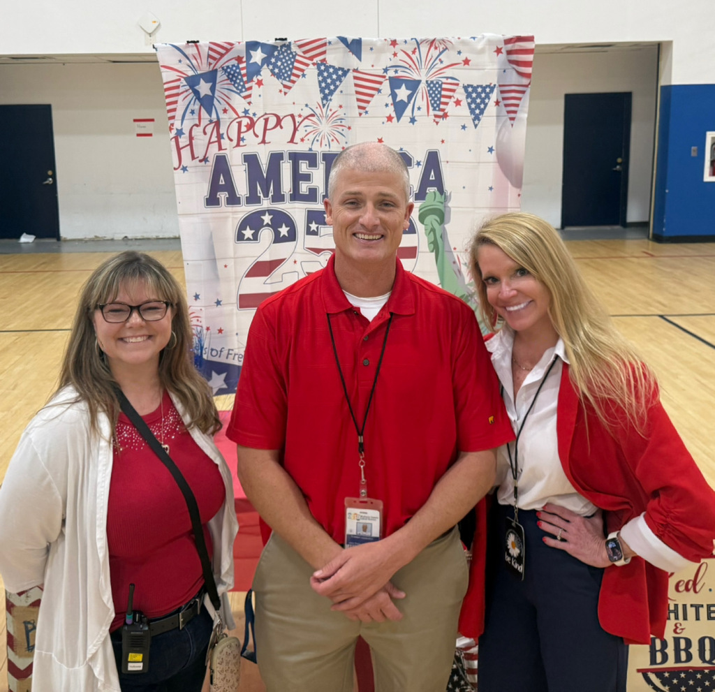 Three staff members pose in front of an America250 patriotic backdrop in the gym.