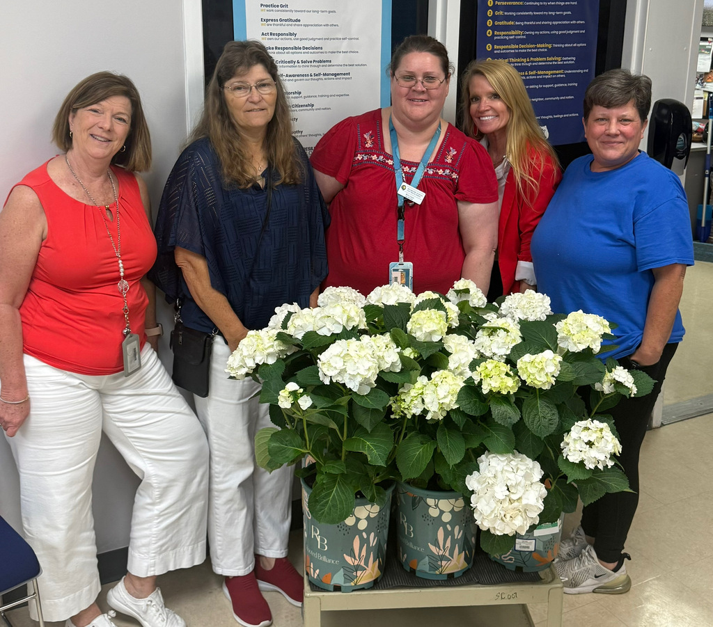 Staff members dressed in red, white and blue pose together beside white hydrangea plants during the school celebration.
