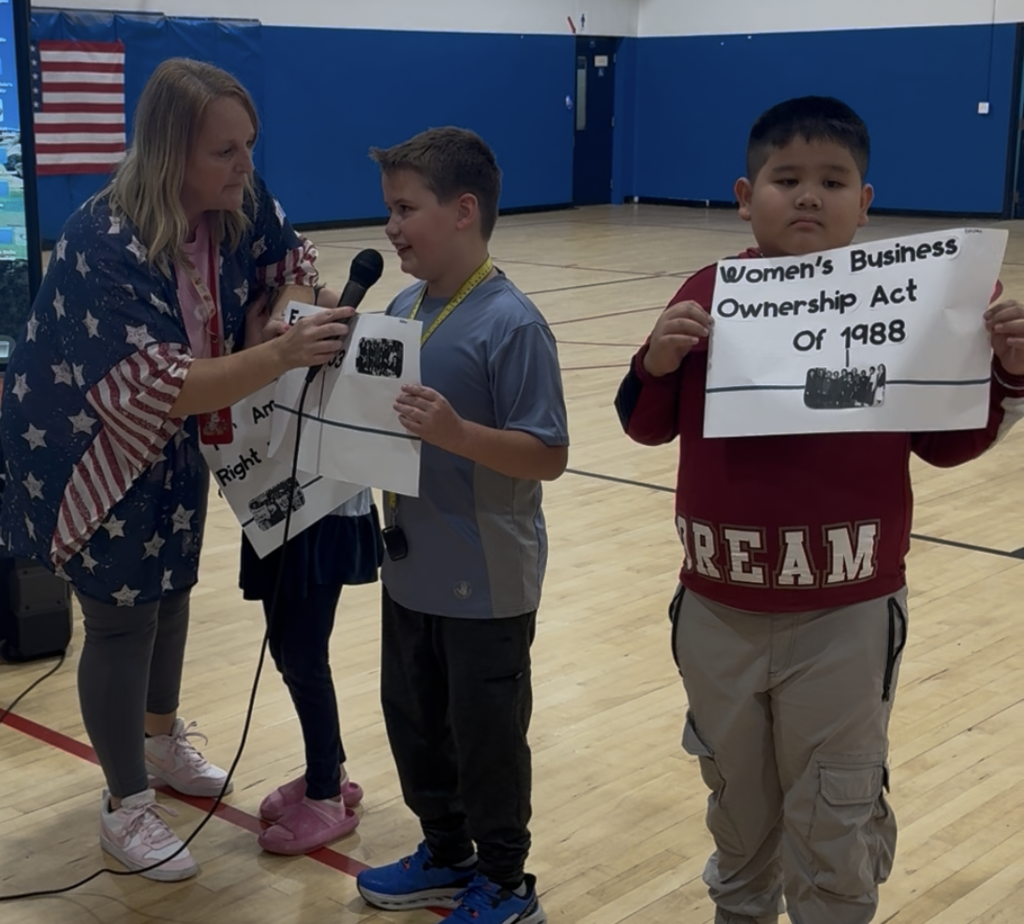 Staff member wearing patriotic colors holds a microphone while students present posters about important events in women’s rights history during the America250 celebration in the school gym.