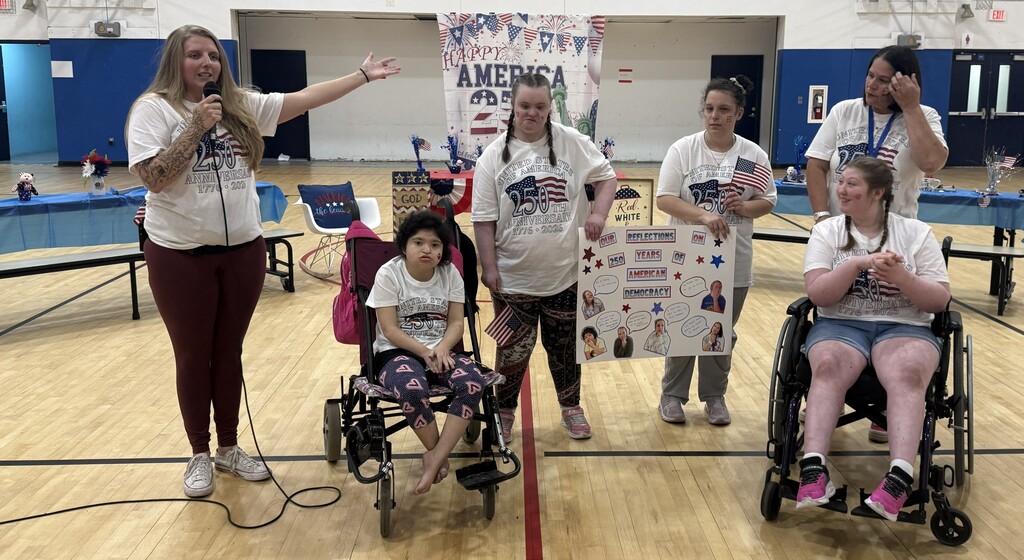 Students and staff wearing patriotic America250 shirts present a poster and speak into a microphone during Richbourg School’s America250 celebration in the gymnasium.