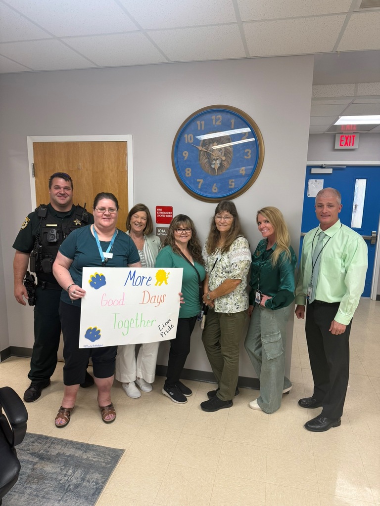 Staff members and a school resource officer wearing green pose inside Richbourg School holding a sign that reads “More Good Days Together” in recognition of Mental Health Awareness Month.