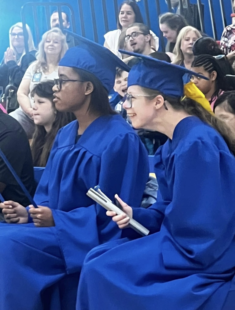 Two graduating seniors wearing blue caps and gowns sit together during the Richbourg Class of 2026 graduation ceremony while family members and guests watch from the bleachers.