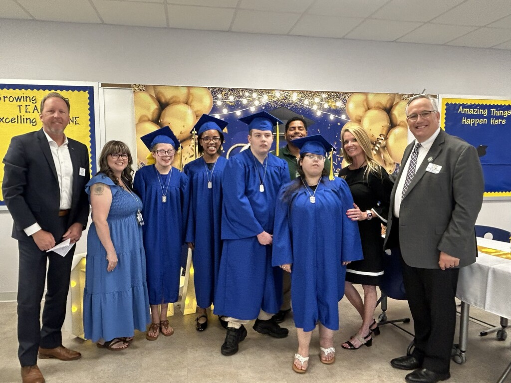 Graduates, school board members Mr. Hiney and Mr. Bryant, Ms. Holloway, the school counselor, and Mrs. Windham, the Assistant Principal, gather together in front of a decorated graduation backdrop during the Class of 2026 celebration.