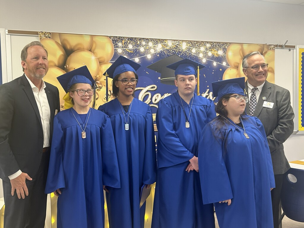 Graduating seniors in blue caps and gowns pose with school and district leaders in front of a graduation backdrop.
