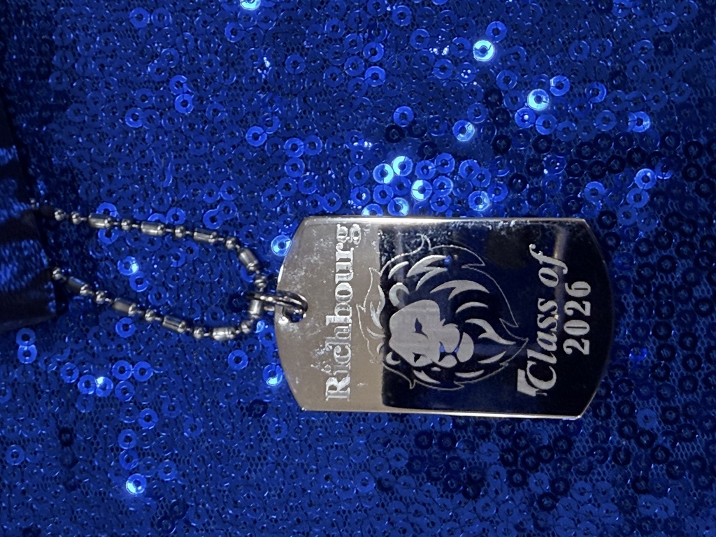 Close-up of a commemorative graduation necklace created by Joey the Jeweler featuring the Richbourg lion logo and engraved with “Richbourg Class of 2026,” displayed on a blue sequined table cloth.