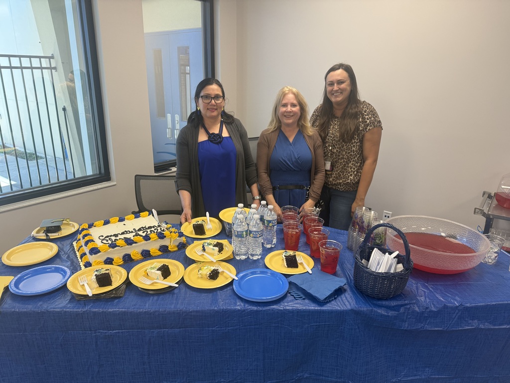 Staff members stand behind a refreshment table with graduation cake, drinks, and desserts during the graduation reception.