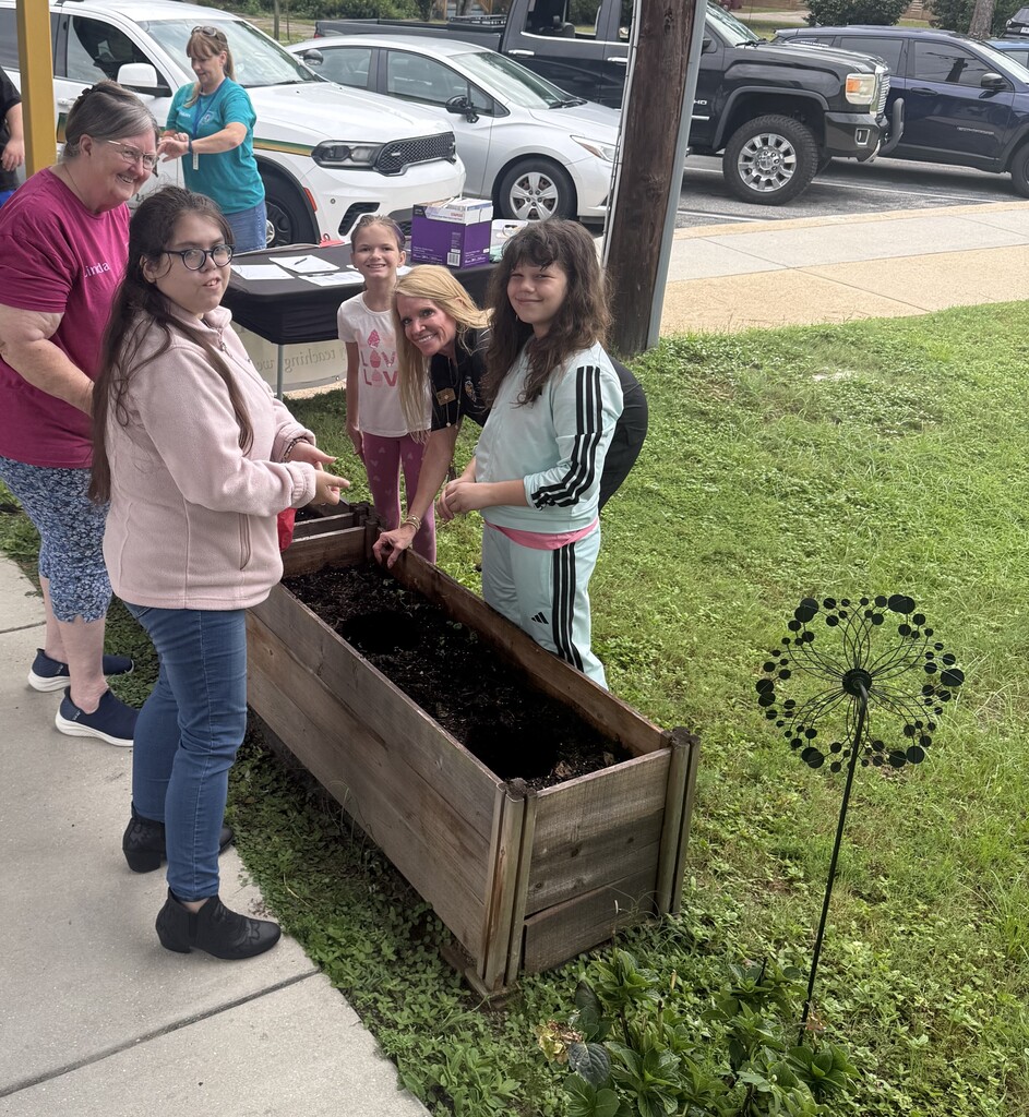 Richbourg students, Mrs. Windham, and other adults smile together while working around a raised garden bed during a hands-on planting activity on the school campus.
