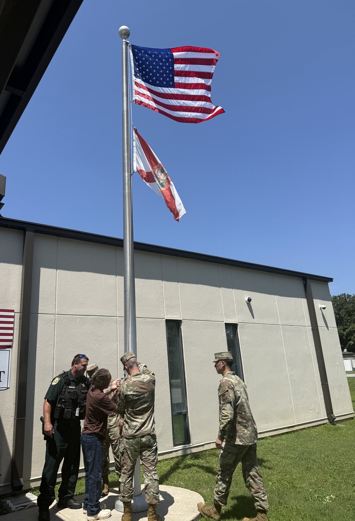 “A Richbourg student, the school resource officer, and members of the Eglin Air Force Base Honor Guard work together at the school flagpole beneath the United States and Florida flags.”