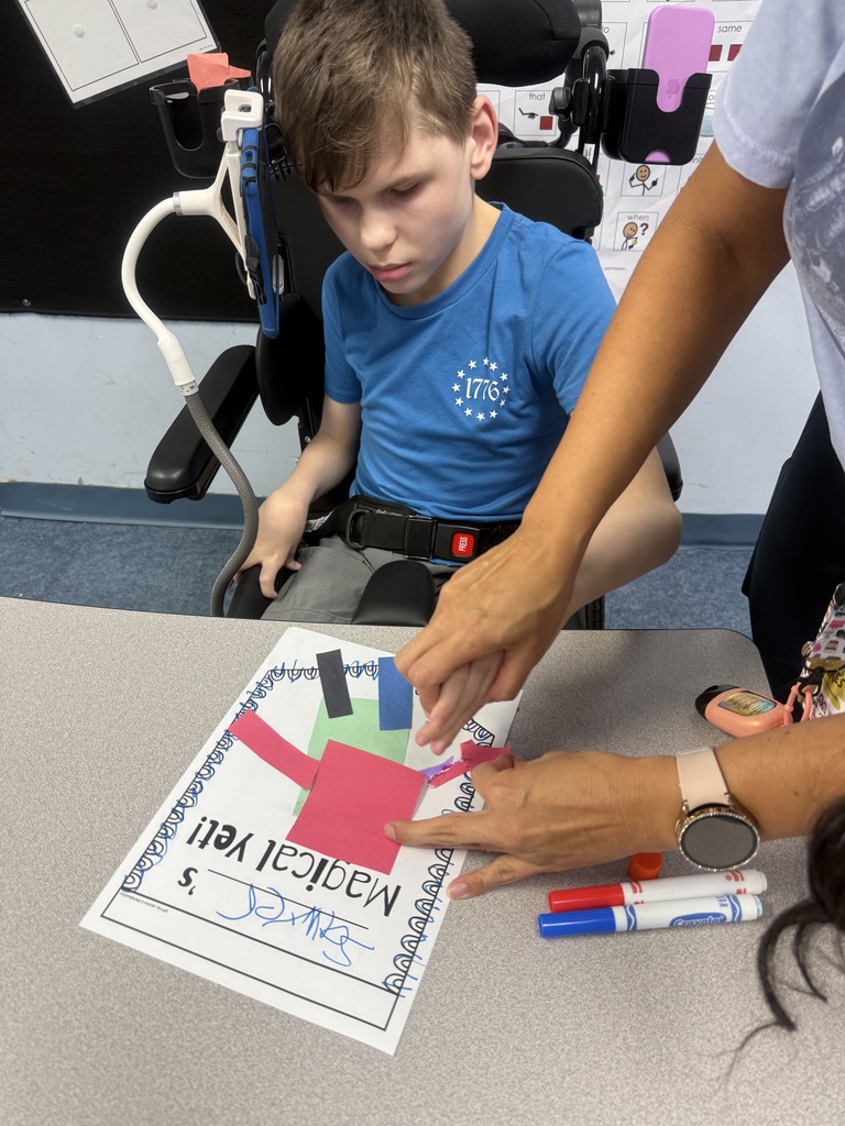 A student works on a “Magical Yet” art project at a classroom table with assistance from an adult. The project is made from colorful paper shapes attached to a worksheet inspired by the book The Magical Yet, reinforcing lessons about perseverance and growth.