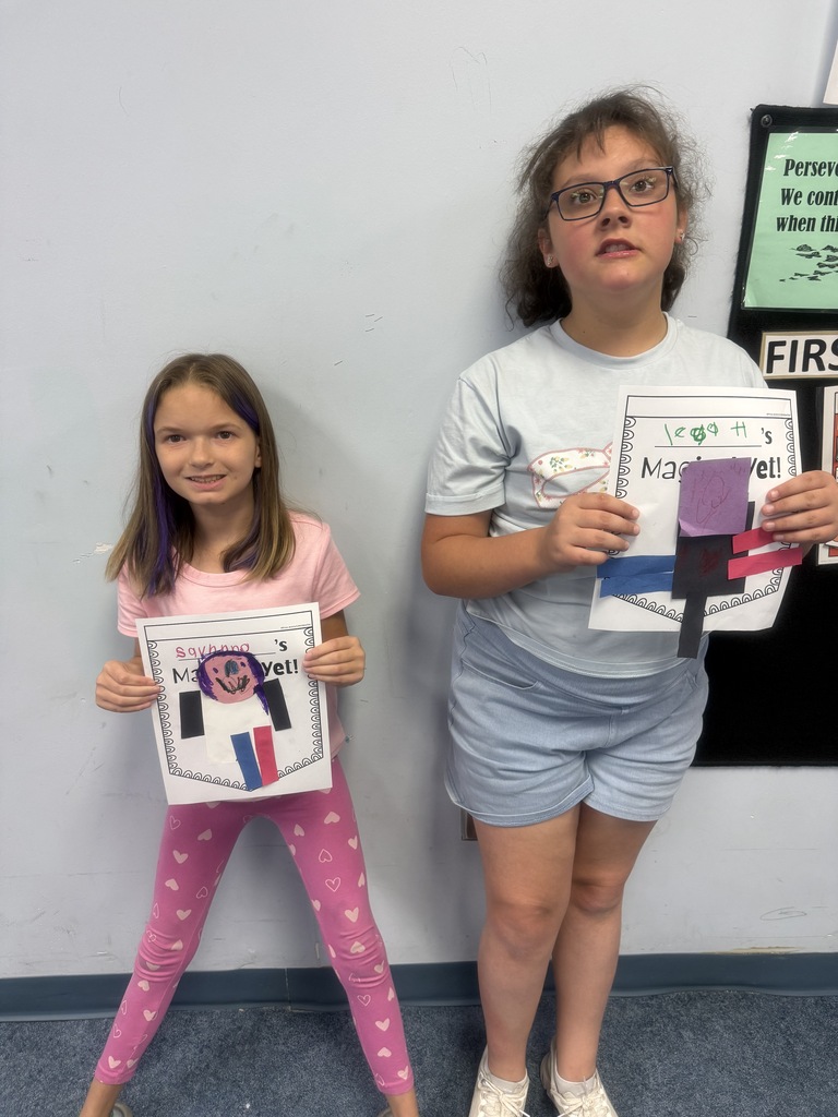 Two students stand in a classroom holding completed “Magical Yet” art projects. The projects feature colorful paper shapes arranged into figures on worksheets inspired by the book The Magical Yet and demonstrate learning about perseverance and growth.