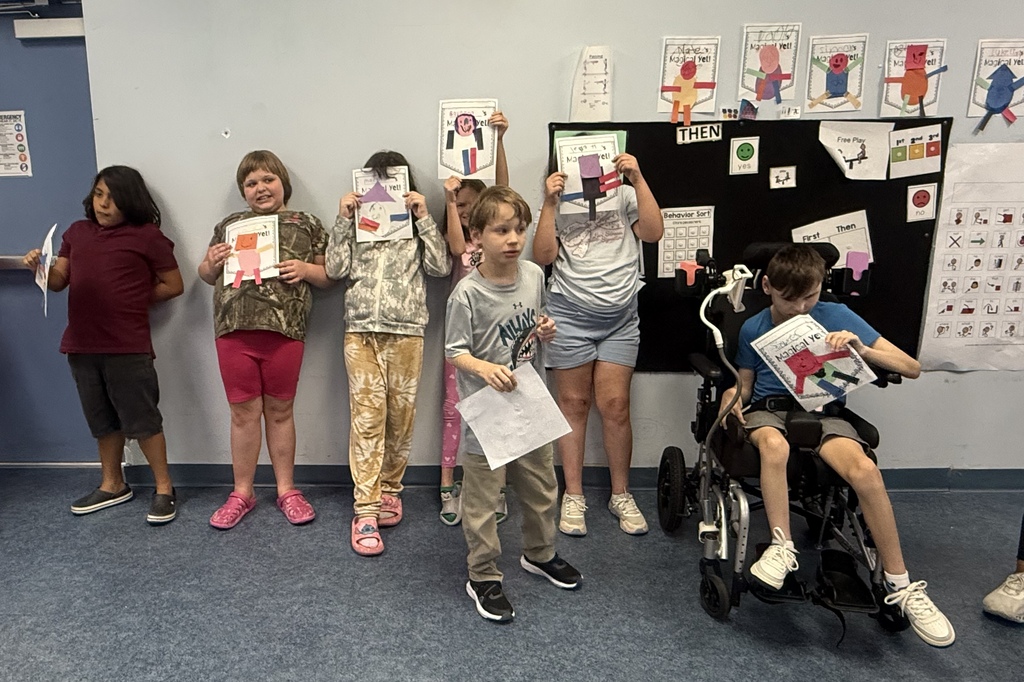 A group of students stands in a classroom displaying completed "Magical Yet!" projects made from colorful paper shapes. Several students hold their projects in front of themselves, while one student stands in the foreground holding a sheet of paper. Another student seated in a wheelchair displays a completed project. Behind the group, a classroom bulletin board features visual supports, including a "First Then" chart, behavior visuals, and communication symbols. Additional student projects are displayed along the wall above the bulletin board. The classroom has blue carpeting and light-colored walls.