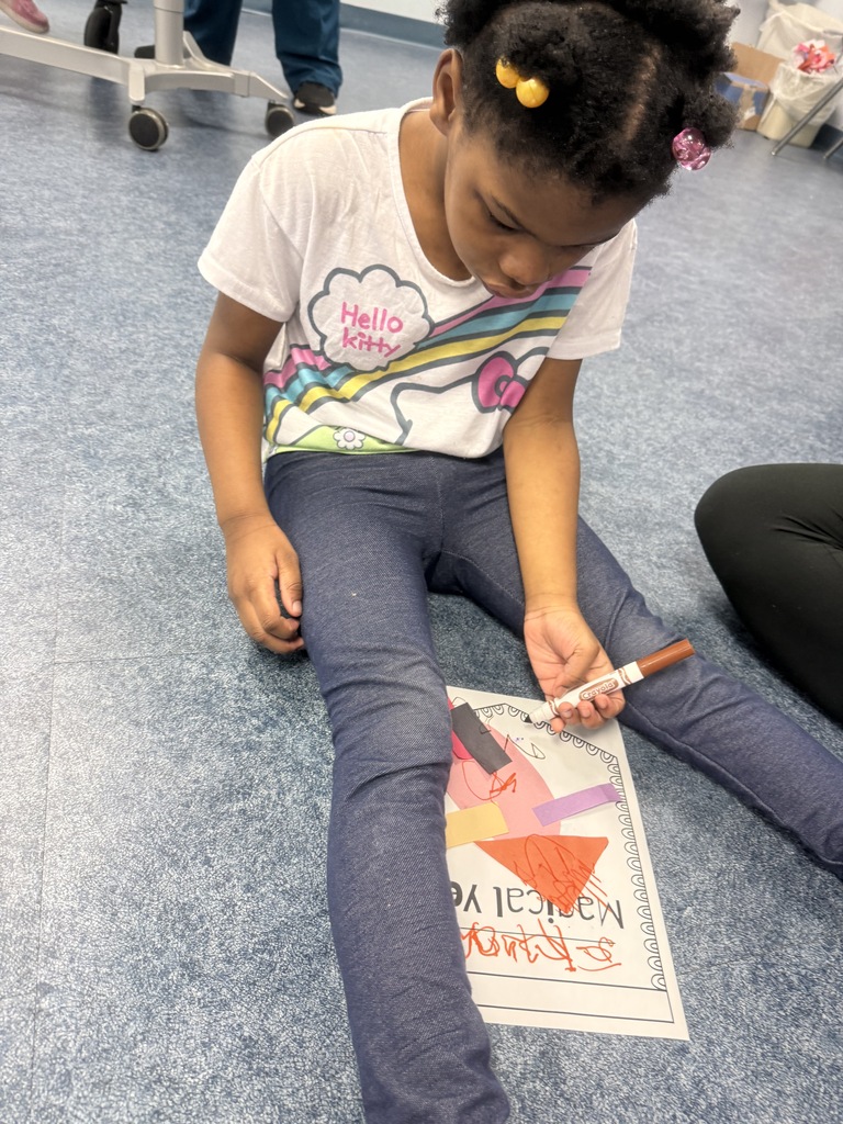 A student sits on a blue classroom floor working on a "Magical Yet!" activity. The student holds a brown marker while coloring and decorating a worksheet with colorful paper shapes and drawings. The worksheet rests on the floor between the student's legs. Part of another person seated nearby and classroom furniture are visible in the background. The activity focuses on creativity, fine motor skills, and classroom participation.
