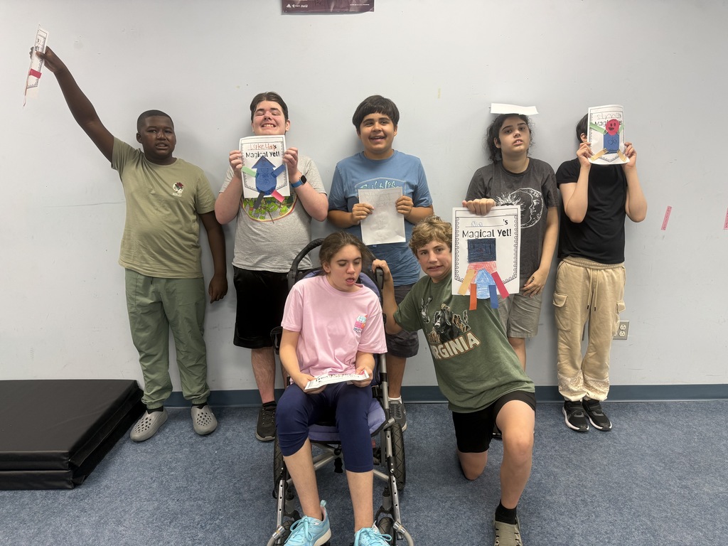 A group of seven students pose together in a classroom against a light-colored wall while displaying completed classroom projects. Several students hold colorful papers labeled “Magical Yet!” featuring hand-crafted designs. One student sits in a wheelchair at the center front holding a paper, while another student kneels beside the wheelchair displaying a project. Other students stand behind them, holding their work for the camera. The classroom has blue flooring and gym mats are visible along the left side of the image.