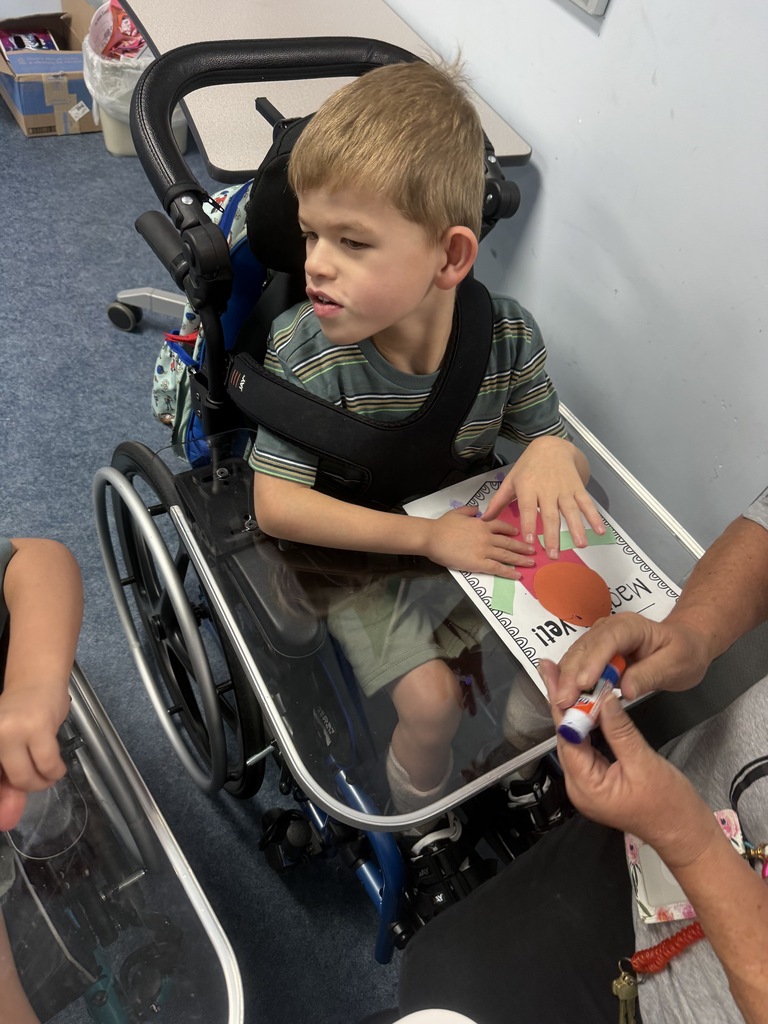 A student seated in a specialized wheelchair participates in a classroom craft activity. The student places colorful paper shapes onto a "Magical Yet!" worksheet positioned on a clear tray attached to the wheelchair. An adult seated nearby assists by holding a glue stick while the student works on the project. The classroom has blue flooring, and another student's arm and workspace are partially visible in the foreground. The activity supports creativity, fine motor development, and classroom engagement.