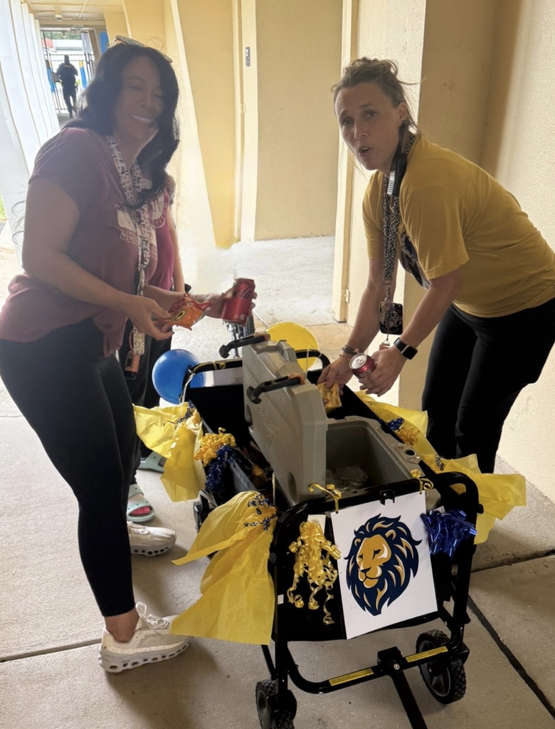 Two staff members stand beside the decorated ROAR Wagon in an outdoor school walkway. The wagon, decorated with blue and gold tissue paper, bows, balloons, and a lion logo, is stocked with snacks and drinks for staff appreciation. The staff members select refreshments from the wagon as it makes its rounds around campus to recognize and thank employees for their dedication to students.