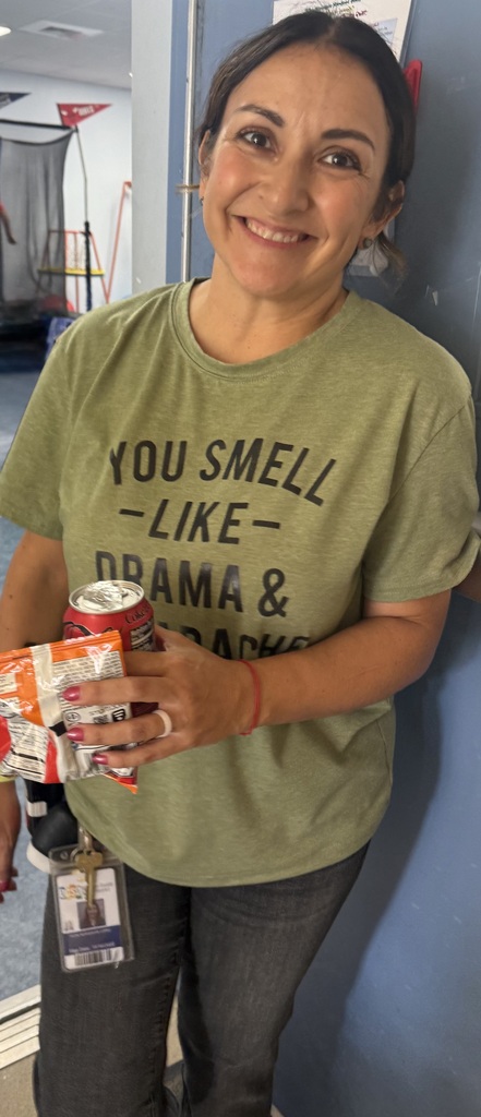 A staff member stands in a classroom doorway holding a bag of chips and a canned drink received from the ROAR Wagon. The staff member wears a green graphic T-shirt, jeans, and an identification badge. Educational equipment and classroom materials are visible in the background. The photo was taken during a Richbourg /Best Chance School staff appreciation event recognizing employees for their dedication and support of students.