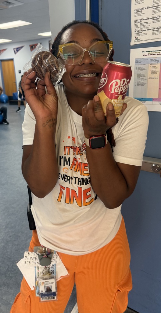 A staff member stands in a school hallway holding a packaged cookie and a can of Dr Pepper & Cream Soda received from the ROAR Wagon. The staff member is wearing glasses, an identification badge, a white graphic T-shirt, and orange pants. Classroom and behavior support information posters are visible on the wall in the background as part of a staff appreciation event at Richbourg/Best Chance School.