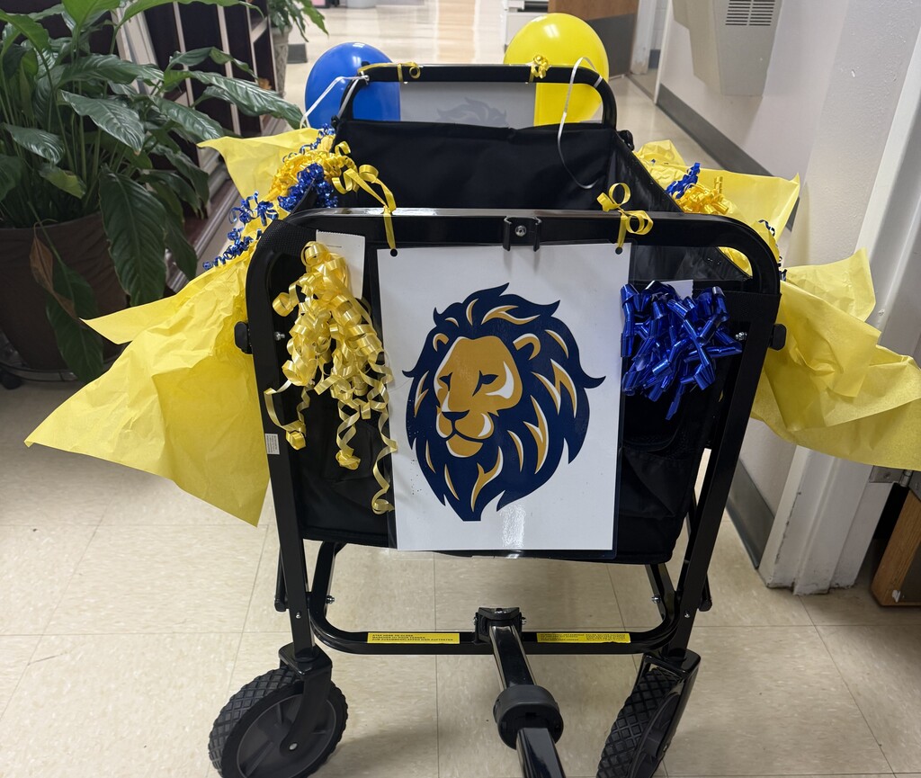 Decorated ROAR Wagon displayed in a school hallway. The black utility wagon features a blue and gold lion logo on the front and is adorned with yellow tissue paper, blue and gold curling ribbon, bows, and balloons. The wagon is used to deliver appreciation treats and recognition to staff members at Richbourg School.