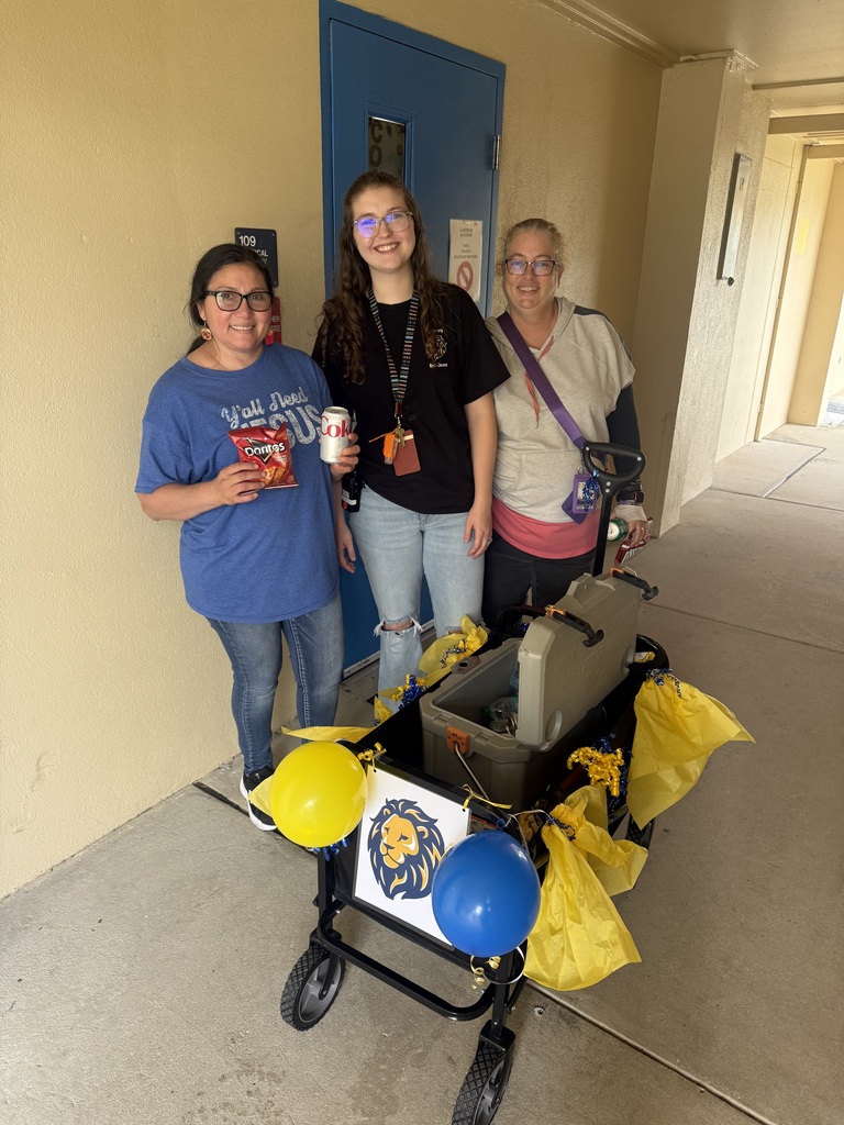 Three staff members stand beside the ROAR Wagon in a covered outdoor walkway at Richbourg School. The decorated wagon features blue and gold balloons, tissue paper, and a lion logo on the front. One staff member holds a bag of chips, another holds a canned drink, and the wagon contains a cooler with beverages and refreshments. The photo was taken during a staff appreciation event recognizing employees for their dedication and support of students