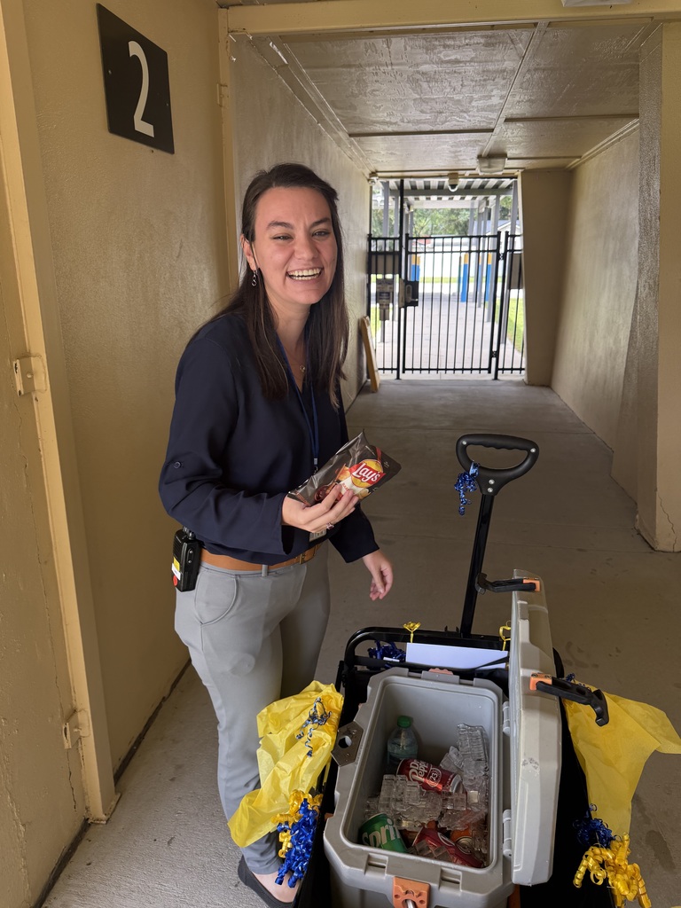 A staff member stands beside the ROAR Wagon in a covered outdoor walkway at Richbourg School. The decorated wagon, adorned with blue and gold tissue paper and ribbons, contains a cooler filled with ice, bottled water, and canned drinks. The staff member holds a bag of chips selected from the wagon during a staff appreciation event recognizing employees for their dedication and service to students.