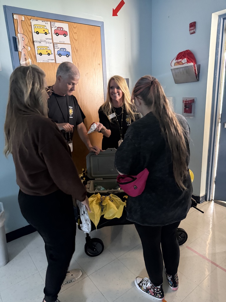 Several staff members gather around the ROAR Wagon inside a classroom at Richbourg School. The decorated wagon, featuring blue and gold school colors, is filled with appreciation treats as staff members select items during a staff recognition visit. Classroom walls and educational materials are visible in the background.