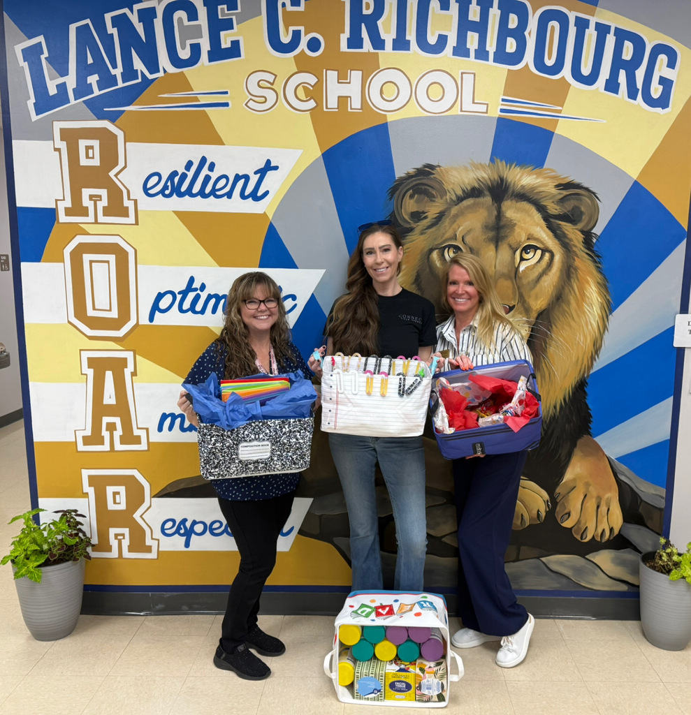 Three adults stand in front of a colorful Richbourg School ROAR mural featuring a large lion. The group displays donated school supplies, including notebooks, folders, markers, and classroom materials contained in baskets and bags. Additional supplies are arranged in a tote on the floor. The donation supports student learning and classroom needs at Richbourg School.