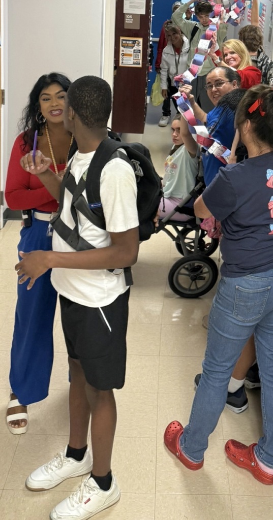 A school hallway activity features students, staff, and families standing along both sides of the corridor holding a long red, white, and blue paper chain. A student wearing a backpack walks through the center of the hallway while participants display the chain, which appears to contain handwritten messages. The activity takes place near classroom entrances and patriotic decorations, creating a community-focused remembrance and unity event.