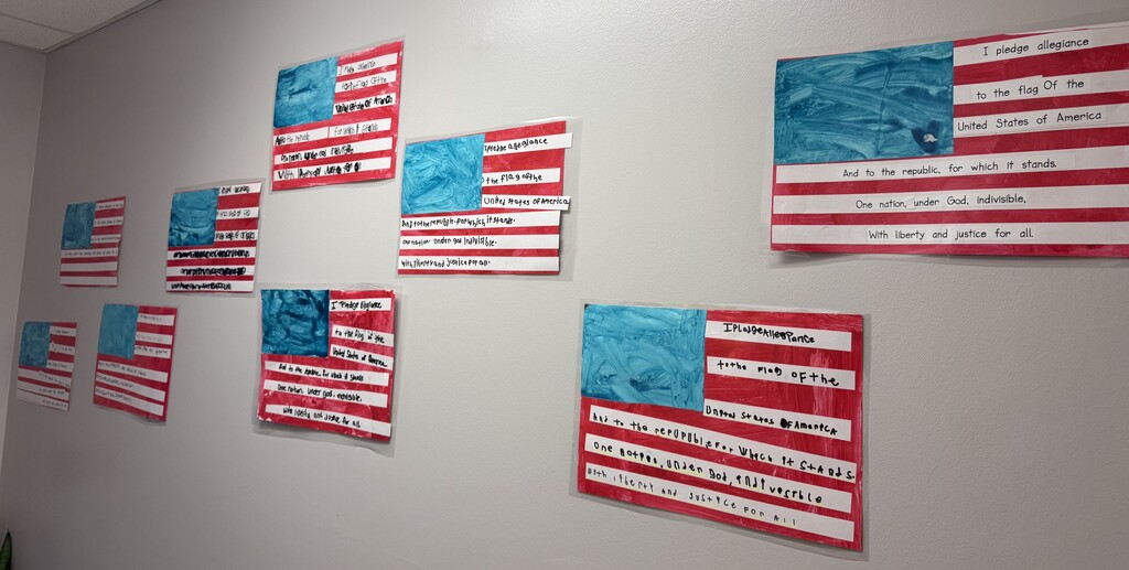 Student-created American flag art projects are displayed on a classroom wall. Each flag is painted with red and white stripes and a blue field, with strips of handwritten text from the Pledge of Allegiance attached across the stripes. Multiple student projects of varying sizes are arranged in a gallery-style display.