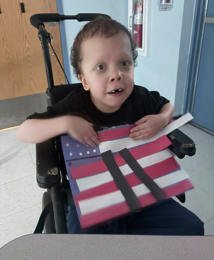 A student seated in a wheelchair displays a handmade September 11 memorial flag craft. The craft includes red and white stripes, a blue field with white stars, and two black rectangular shapes symbolizing the Twin Towers as part of a classroom patriotism lesson.