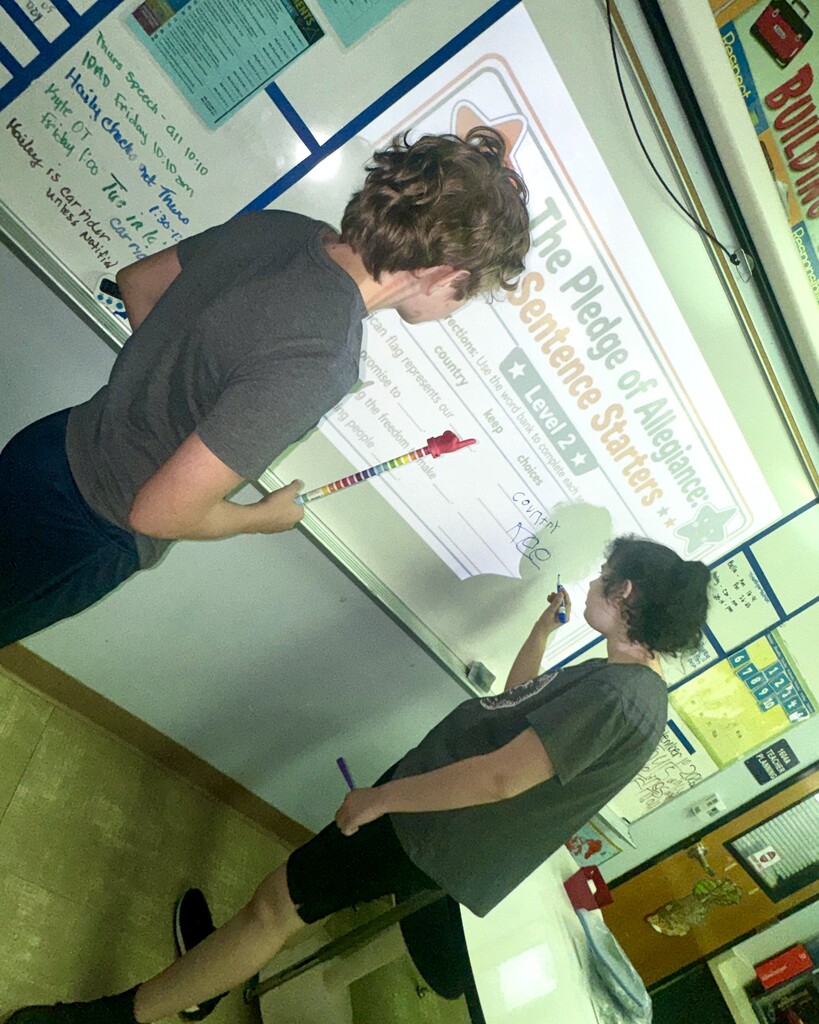 Two students work at an interactive whiteboard displaying a lesson titled “The Pledge of Allegiance Sentence Starters, Level 2.” The students use markers to complete sentences about patriotism and citizenship during a classroom literacy activity.