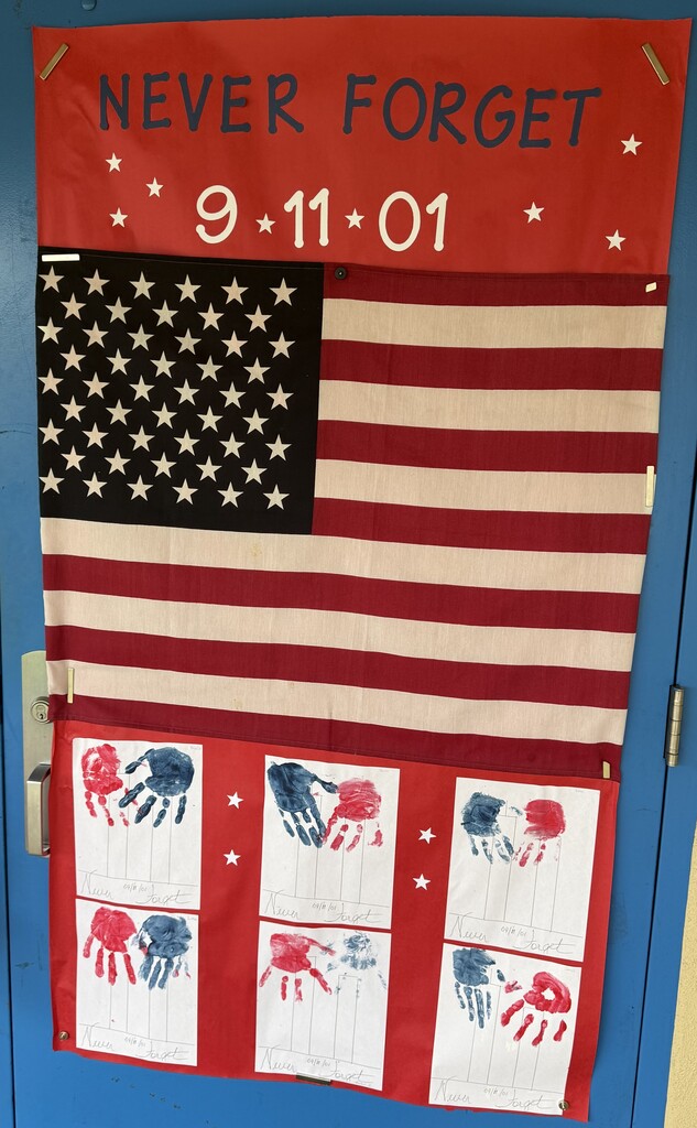 A patriotic classroom display features a large American flag mounted on a blue door beneath a red banner reading “NEVER FORGET 9.11.01.” Student artwork showing red, white, and blue handprints is displayed below the flag as part of a September 11 remembrance activity.