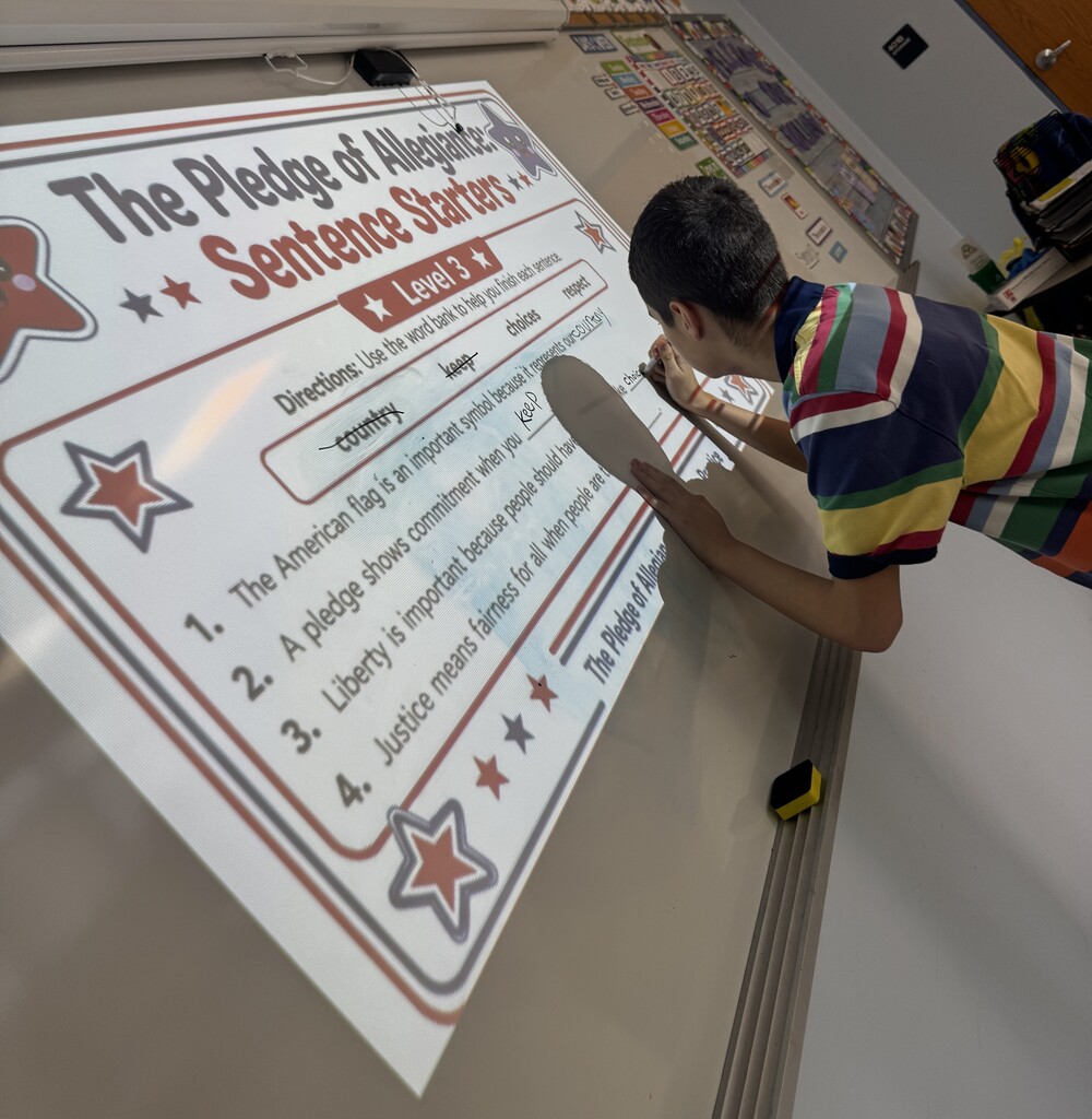 A student writes on an interactive whiteboard displaying a lesson titled “The Pledge of Allegiance Sentence Starters.” The activity includes a word bank and sentence prompts about the American flag, liberty, and justice. The student uses a marker while leaning over the projected lesson in a classroom setting.