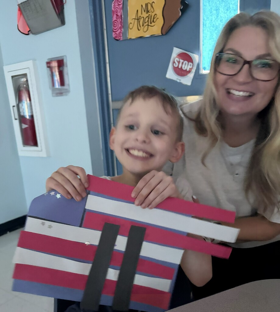 A student displays a handmade September 11 memorial craft alongside a teacher at a classroom table. The craft features an American flag design with red and white stripes, a blue field with stars, and two black rectangular shapes representing the Twin Towers as part of a patriotic remembrance activity.