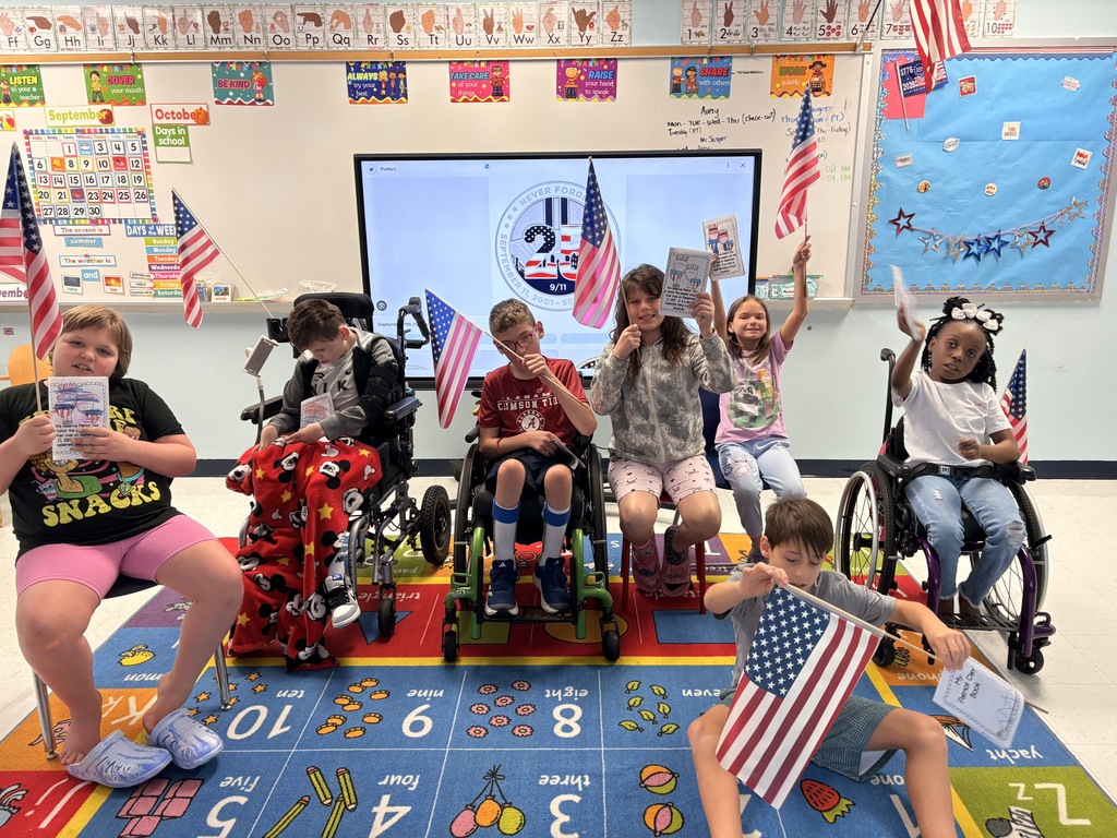 Students in a classroom hold small American flags and completed worksheets during a lesson recognizing September 11. Several students are seated on chairs and adaptive mobility devices around a colorful classroom rug. An interactive display in the background shows a 9/11 remembrance graphic, and patriotic decorations and flags are visible throughout the room.