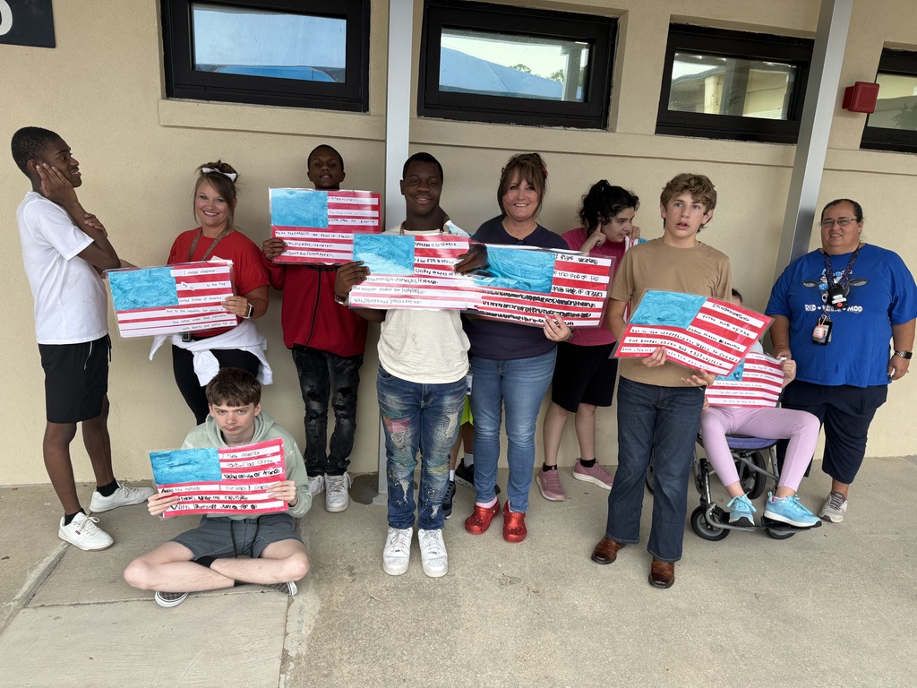 A group of students and staff stand outside a school building holding student-created American flag projects. The artwork features painted blue fields, red and white stripes, and handwritten portions of the Pledge of Allegiance. One student is seated in front of the group displaying a completed project, while others stand in a row showing their work as part of a classroom lesson on patriotism and civic engagement.