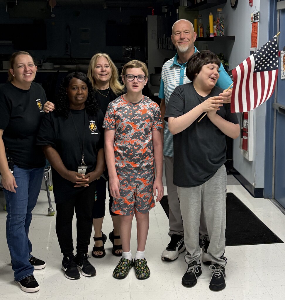 Students and staff stand together in a school hallway near a doorway decorated with an American flag. One student holds a small American flag while the group participates in a patriotic school activity or remembrance event.