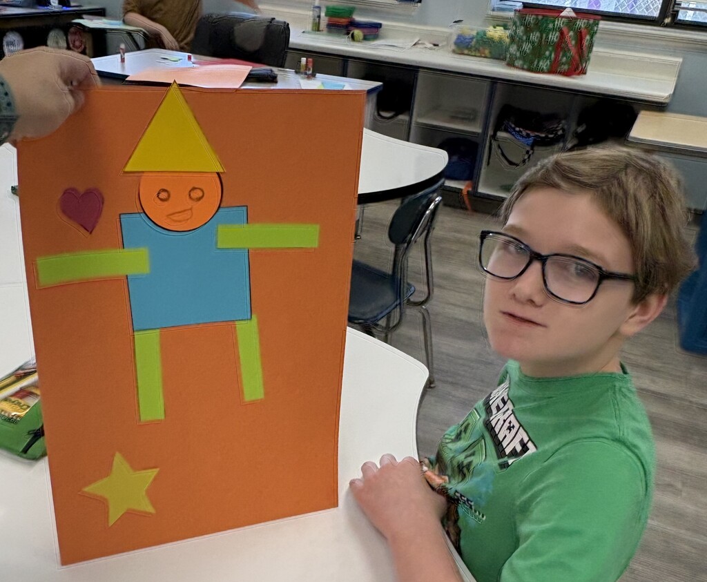 A student displays a classroom art project featuring a person made from colorful geometric shapes on an orange poster board.
