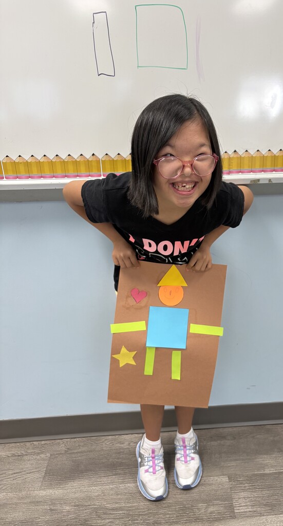 A student displays a geometric shapes poster showing a person made from colorful shapes, along with a heart and star, during a classroom shape recognition lesson.