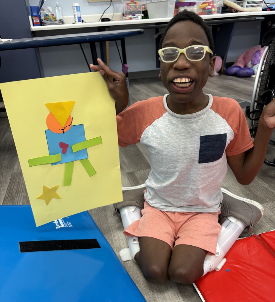 A student holds up a geometric shapes art project showing a person made from colorful shapes on a yellow background during a classroom lesson.