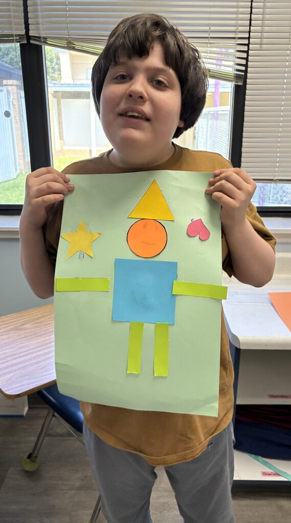 A student displays a geometric shapes artwork showing a person created from colorful shapes on a green poster during a classroom lesson.