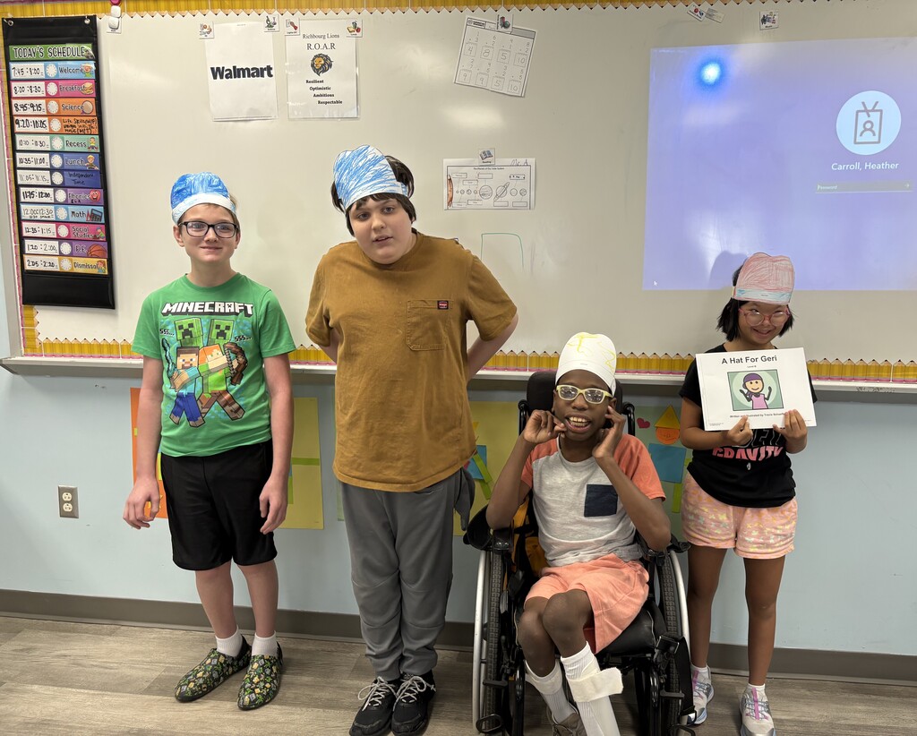 Four students wearing paper hats pose in a classroom during a literacy lesson. One student holds the book A Hat for Geri while standing with classmates in front of a whiteboard.