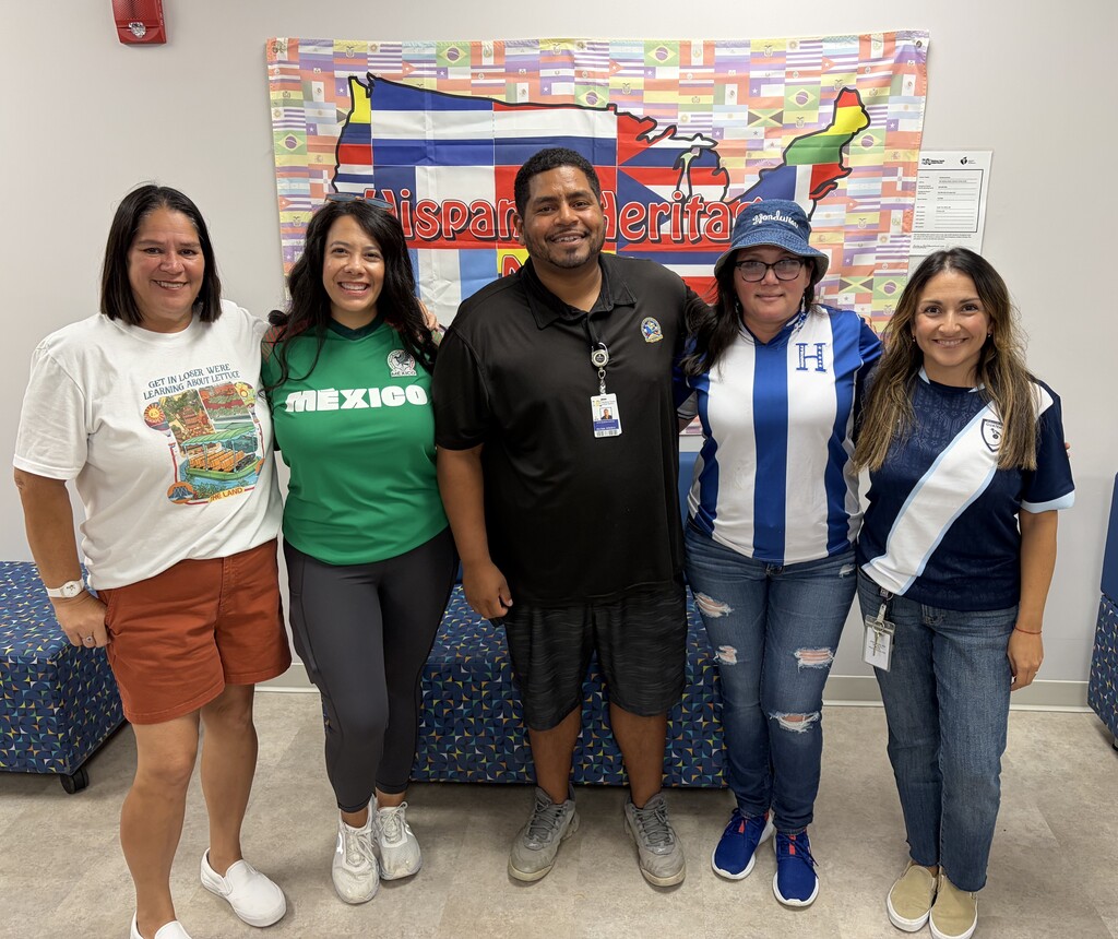 Five staff members pose in front of a Hispanic Heritage Month banner, wearing apparel representing different Hispanic and Latin American countries during a school celebration.
