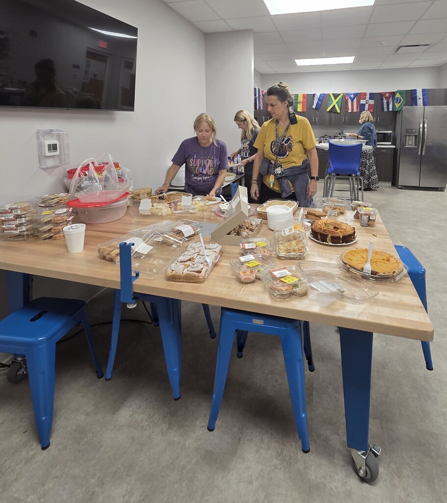 Staff members enjoy a dessert potluck in a school break room, with a table full of pies, pastries, cookies, and other treats.