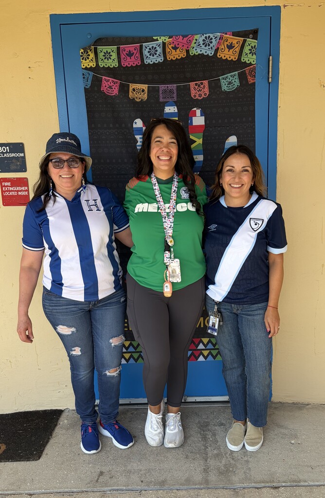 Three staff members stand in front of a decorated doorway during a Hispanic Heritage Month celebration. The doorway display features colorful papel picado banners and flag-themed artwork. The staff members wear shirts representing different Hispanic and Latin American countries and pose together in front of the festive cultural display outside a classroom.