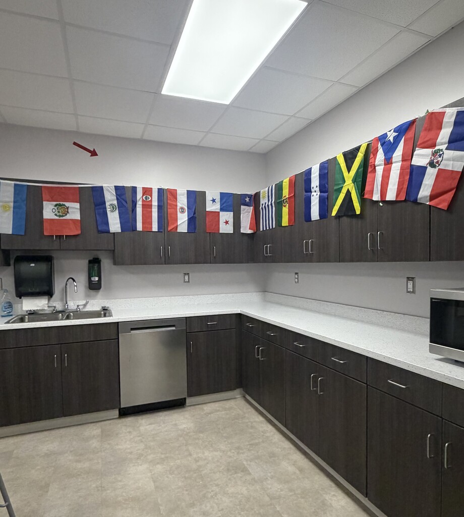 A school staff lounge or kitchenette is decorated for Hispanic Heritage Month with a display of colorful national flags from Hispanic and Latin American countries and regions hanging above the cabinets. The space includes countertops, cabinets, a sink, and a dishwasher, creating a welcoming environment that celebrates the diversity and cultural contributions of Hispanic communities.