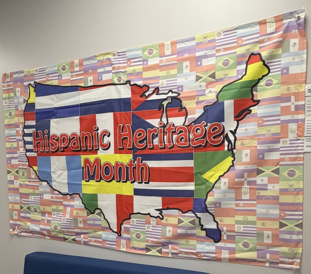 A colorful Hispanic Heritage Month banner hangs on a wall in a school lounge or gathering space. The banner features an outline of the United States filled with the flags of various Hispanic and Latin American countries, set against a background of additional national flags. Large text across the center reads “Hispanic Heritage Month,” celebrating the cultures, histories, and contributions of Hispanic communities.