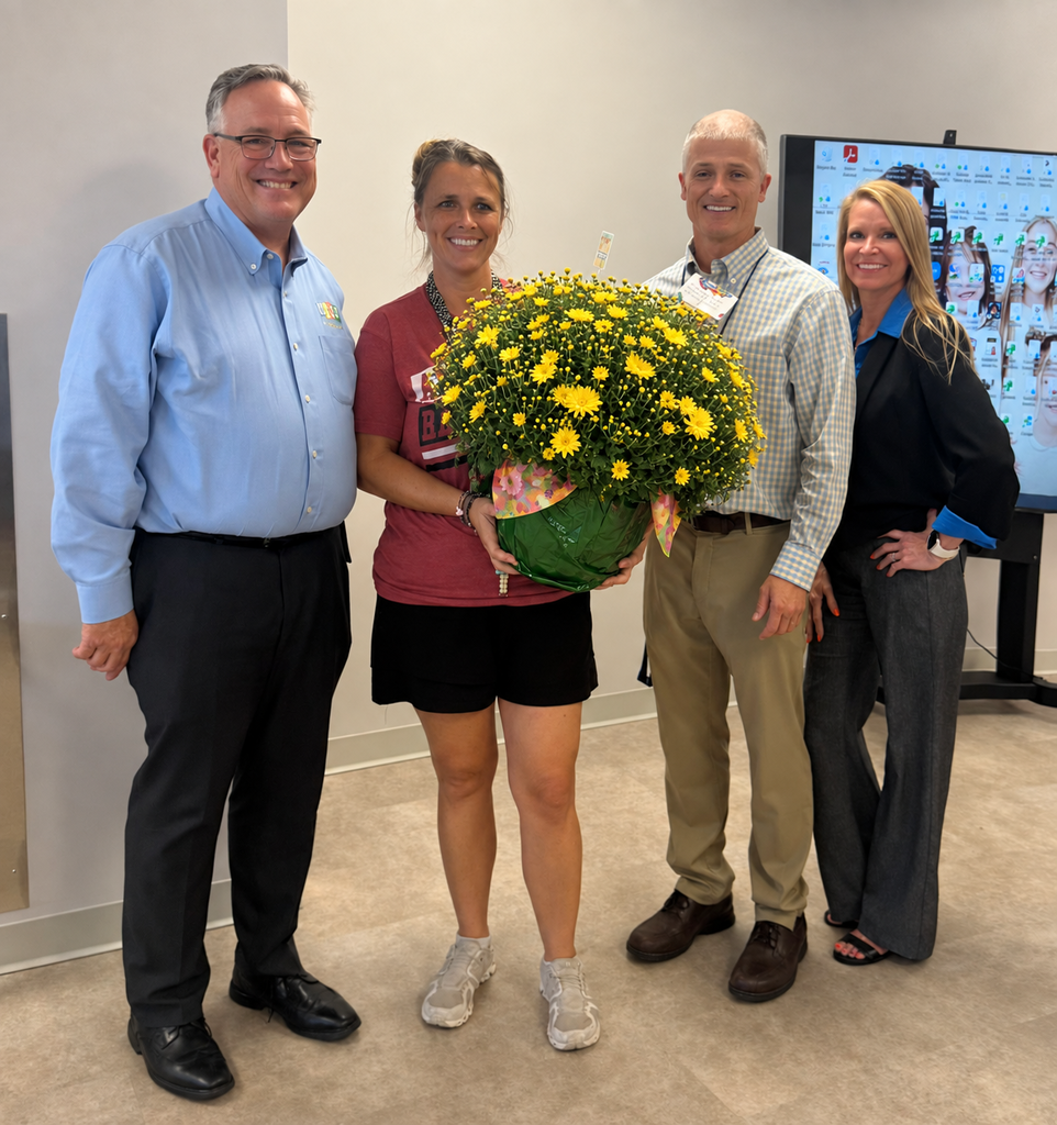 School Board Member Tim Bryant, Richbourg Teacher of the Year Ashley Frost, Principal Mr. Harvell, and Assistant Principal Mrs. Windham stand together indoors, smiling for a photo. Ashley Frost stands in the center holding a large arrangement of bright yellow flowers presented in recognition of her selection as Richbourg’s Teacher of the Year.