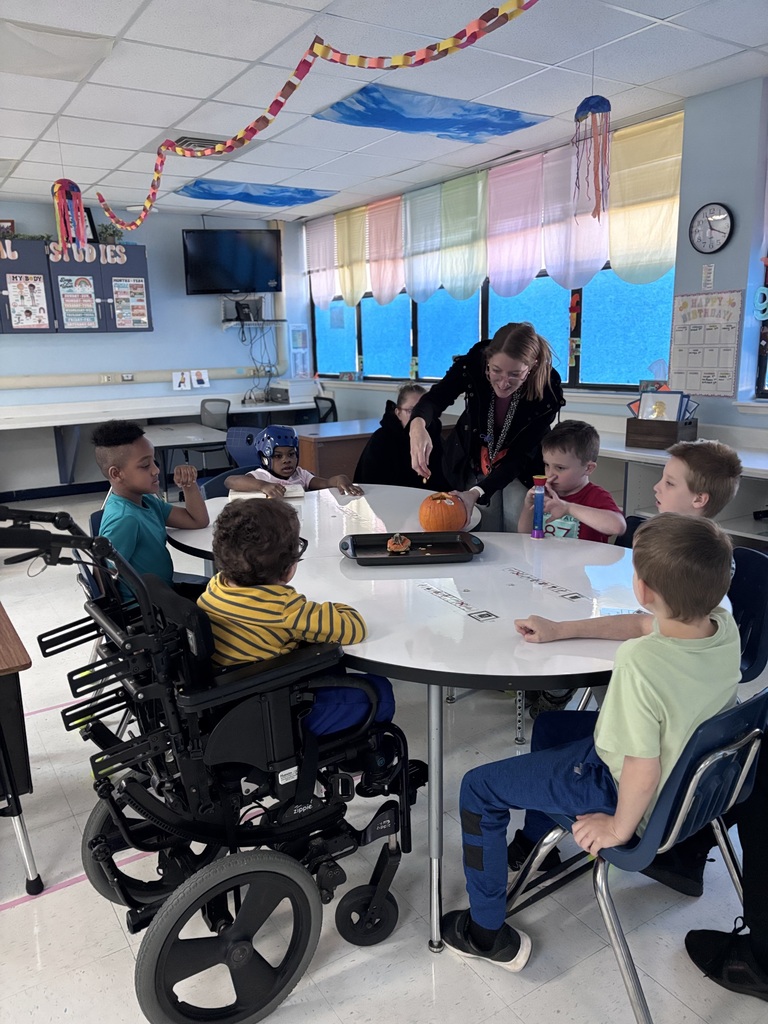 Several students sit around a round classroom table while an educator leads a pumpkin exploration activity. Students engage in hands-on sensory learning by observing and touching a pumpkin. The classroom includes accessible seating and adaptive equipment, supporting participation by all learners. Colorful decorations and educational materials are visible throughout the classroom.