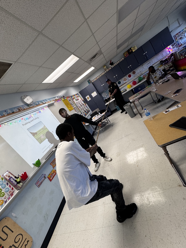 Students participate in an interactive science lesson on push and pull by using a rope stretched across the classroom. One student pulls on the rope while another student helps demonstrate the force being applied. A school resource officer stands at the opposite end of the rope participating in the activity. A classroom presentation titled "Pushing and Pulling" is displayed on the board, connecting the hands-on demonstration to the science concept being taught.