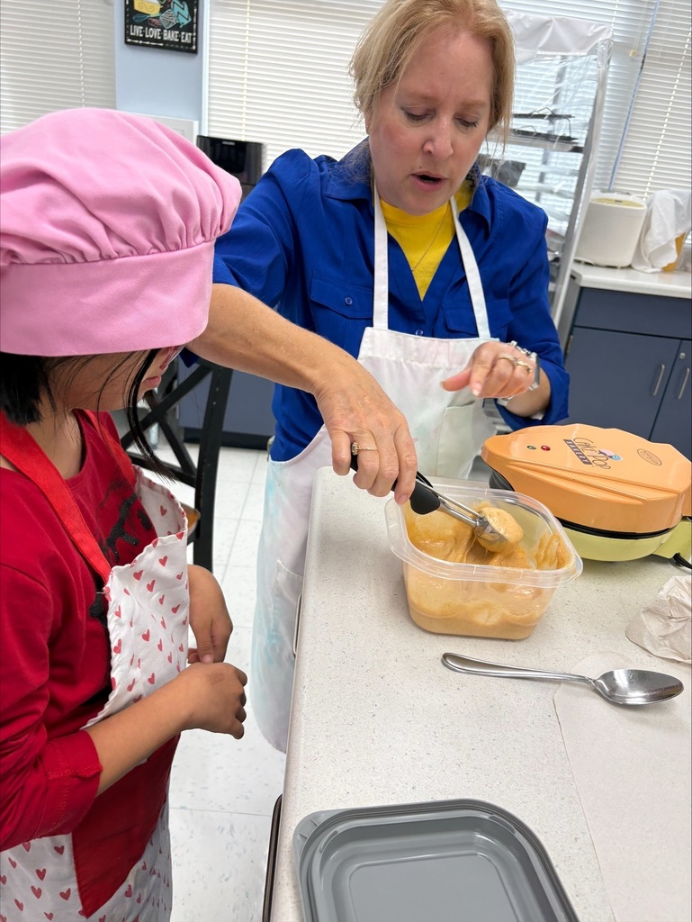 A student wearing a chef hat and apron learns how to make cake pops while an educator demonstrates the next step in the recipe during a hands-on cooking lesson.